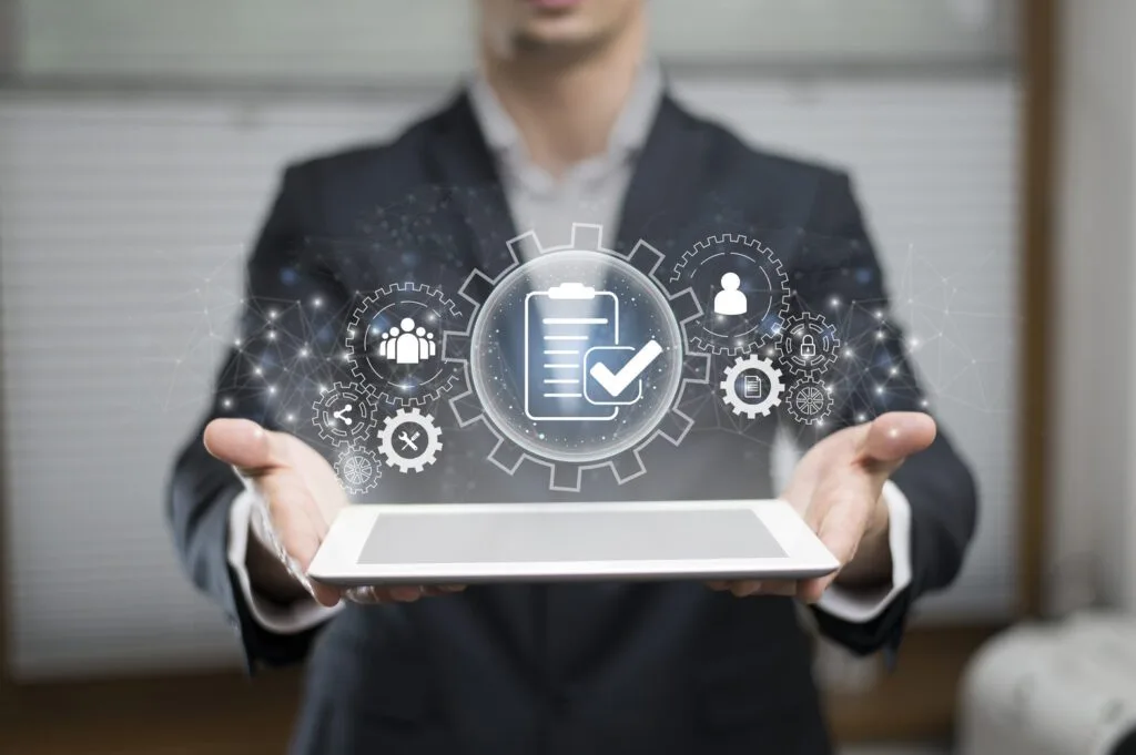 A person in a suit holds a tablet with a holographic interface above it. The hologram features a digital checklist with a checkmark, surrounded by gear icons representing settings and connectivity.
