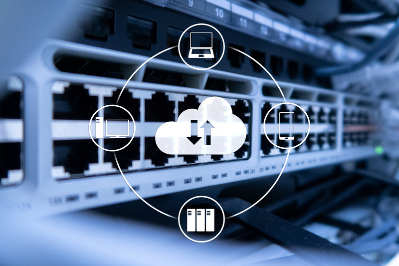 The image shows a network infrastructure, likely part of a server room or data center, with Ethernet ports and cables connected to networking hardware, such as switches or routers. Overlaid on the image is a graphic depicting cloud computing, featuring a central cloud icon with arrows for data transfer (upload and download). Surrounding the cloud are icons representing different devices and systems: a desktop computer, a laptop, a smartphone, and servers, illustrating the concept of interconnected devices and cloud storage or services.