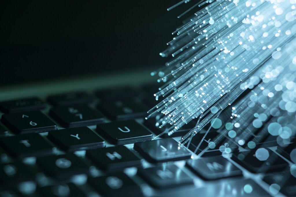 Close-up image of a laptop keyboard illuminated by strands of blue fiber optic cables. The glowing fibers create a bokeh effect, symbolizing high-speed data transmission, technology, and digital connectivity.