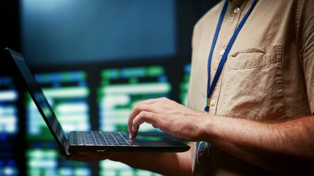 A close-up of a person typing on a laptop in a server room or control center, with large monitors displaying data in the blurred background. The individual wears a beige shirt and a blue lanyard, suggesting a professional, possibly in IT or network security, monitoring or managing server operations.