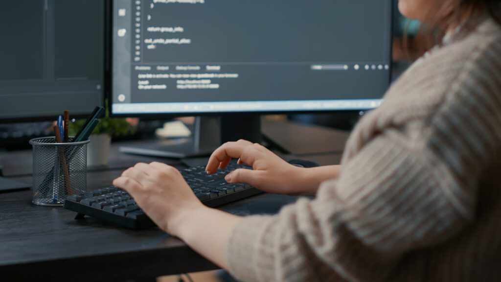 The image shows a person working at a desk, typing on a keyboard, with a focus on their hands. The individual is writing or editing code displayed on two monitors in front of them. The screen content suggests software development or programming, featuring lines of code in a dark-themed code editor. A pen holder with various writing instruments is also visible on the desk, adding a casual office or home office setting to the scene.