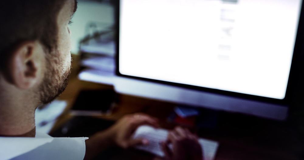 A person with short hair and a beard is working at a computer desk. The individuals hands are on a keyboard and a large monitor displays a bright screen. The environment is dimly lit, with various papers and items on the desk.