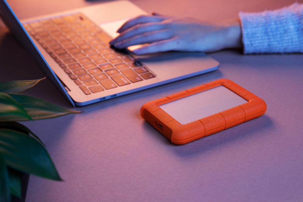 A person using a laptop on a desk next to an orange external hard drive. Soft lighting and a plant in the foreground create a relaxed atmosphere.