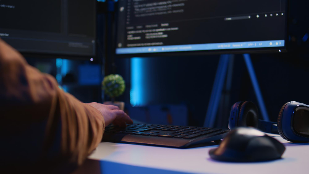 A person types on a keyboard in a dimly lit room with two monitors displaying code. A small plant and headphones are on the desk, creating a focused programming atmosphere.