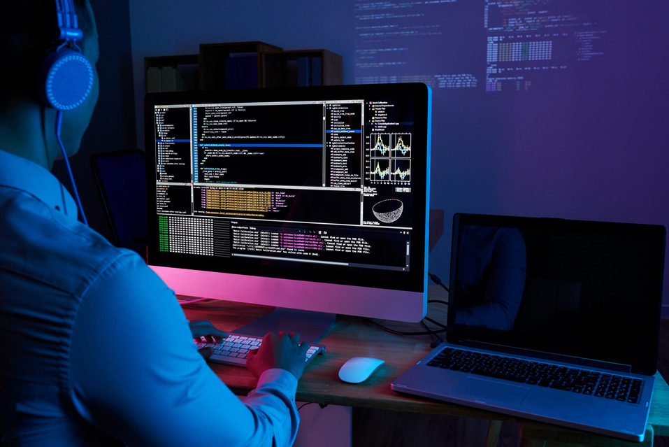 A person wearing headphones coding on a desktop computer in a dimly lit room.