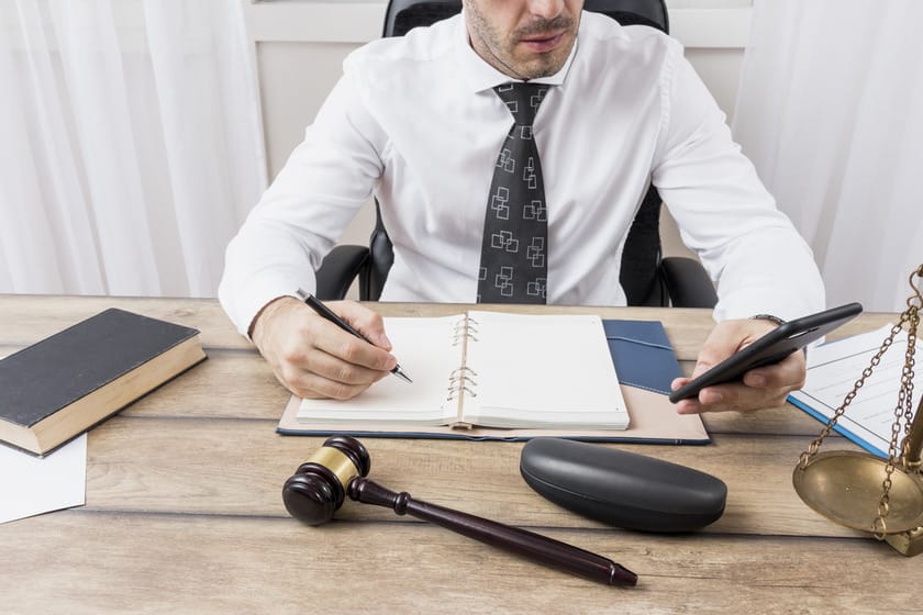 The image shows a man in a professional setting, likely a lawyer, seated at a desk. He's dressed in a white shirt and patterned tie, holding a smartphone in one hand and writing in a notebook with the other.